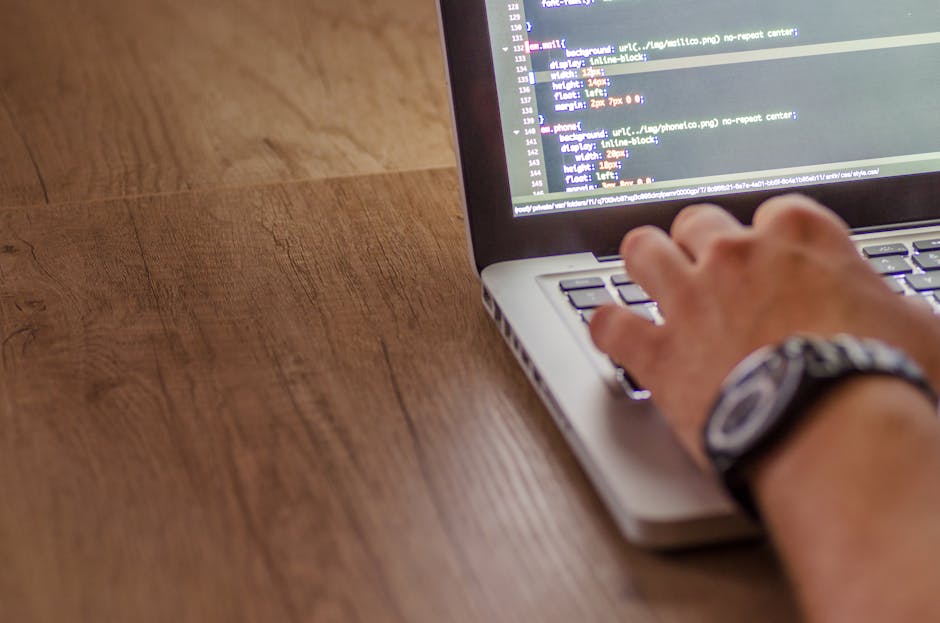 Close-up of a person's hands operating a laptop on a dark table indoors, with the screen displaying lines of code in a dark-themed integrated development environment. The individual appears to be work