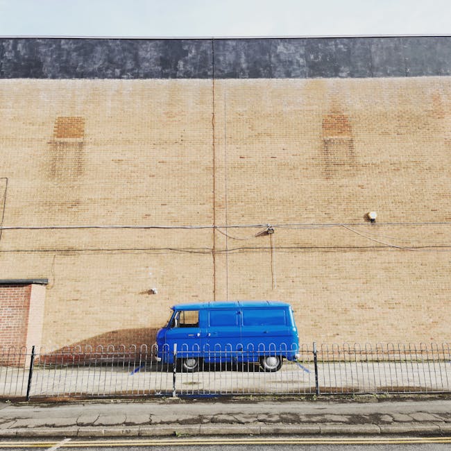 A bright blue vintage van parked on a city street in front of a large brick building with a plain beige and dark gray exterior wall. The van is positioned behind a black metal fence and cast in direct