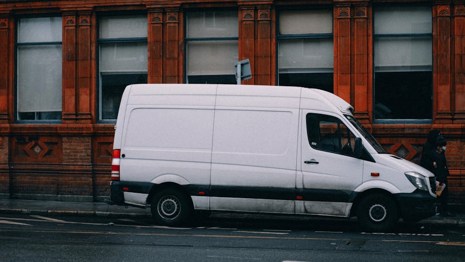 A bright blue vintage van parked on a city street in front of a large brick building with a plain beige and dark gray exterior wall. The van is positioned behind a black metal fence and cast in direct